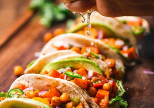 The image shows a hand squeezing lime over colorful tacos filled with chickpeas, vegetables, avocado, and cilantro on a wooden board.