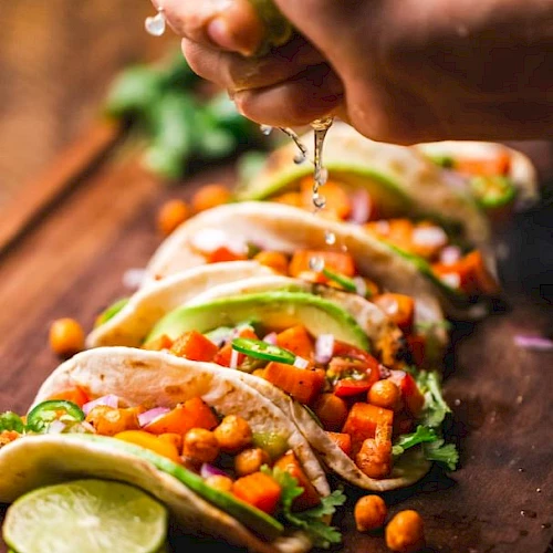The image shows a hand squeezing lime over colorful tacos filled with chickpeas, vegetables, avocado, and cilantro on a wooden board.
