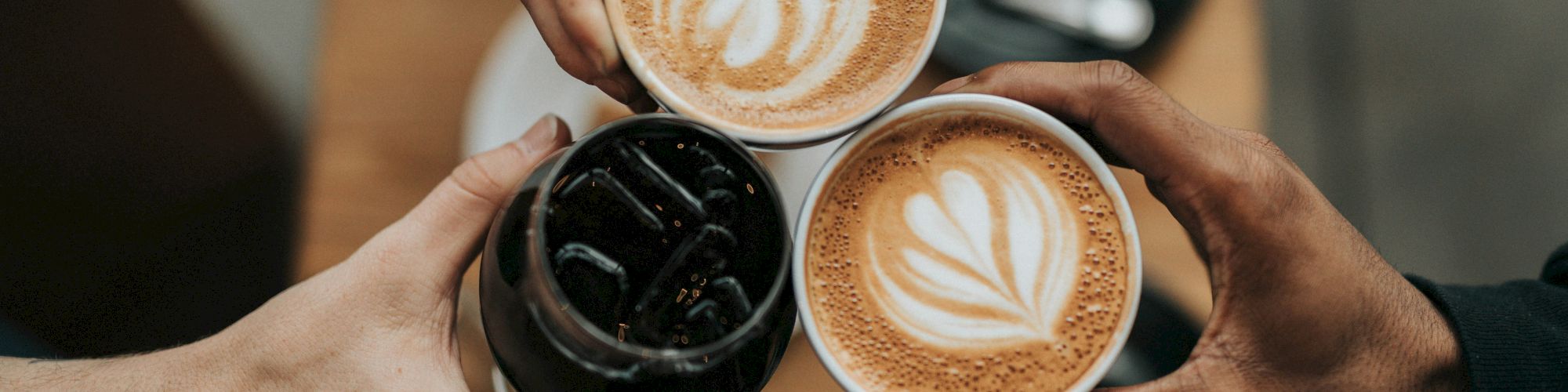 Three hands holding drinks, two lattes with latte art, and one iced beverage, above a wooden table with empty saucers and spoons.
