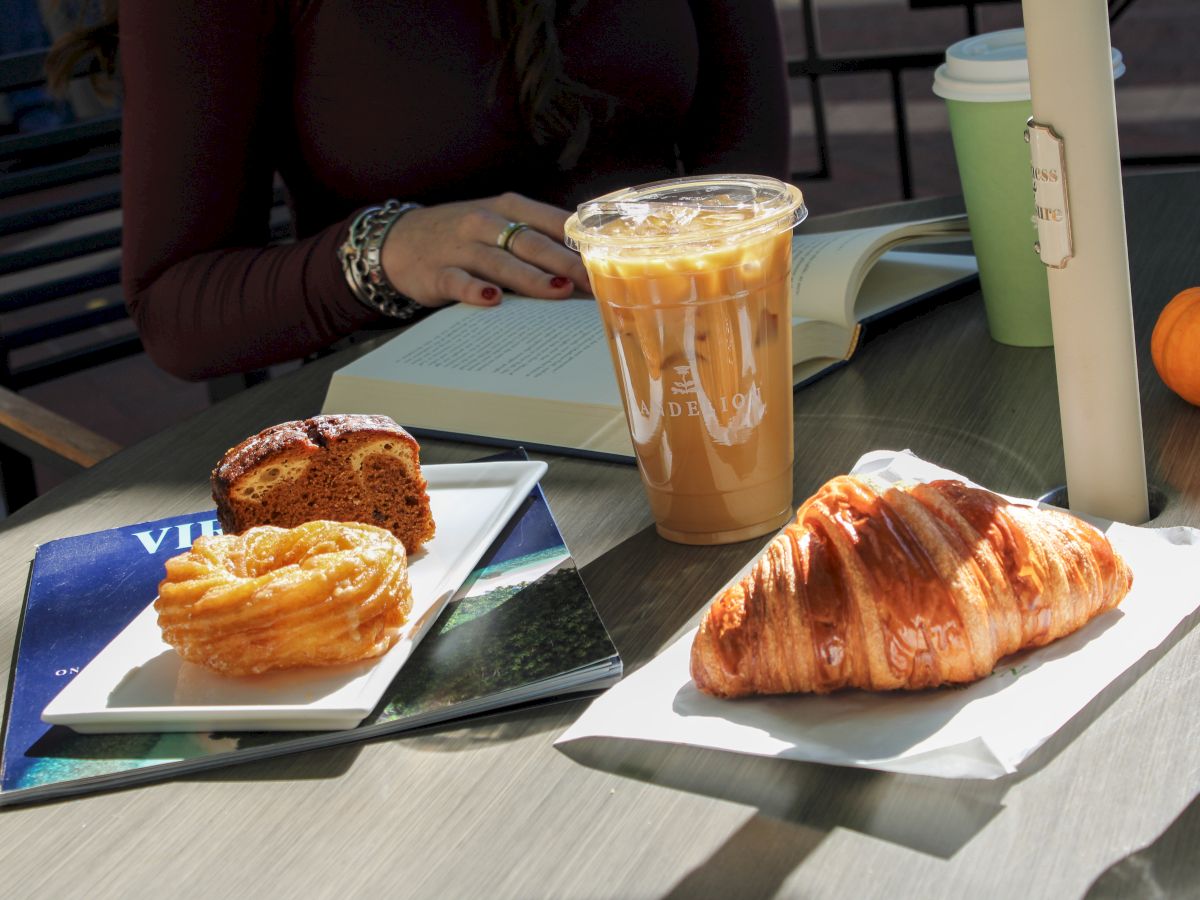 The image shows pastries on plates, an iced coffee, a person reading, and a green cup on a table outdoors.