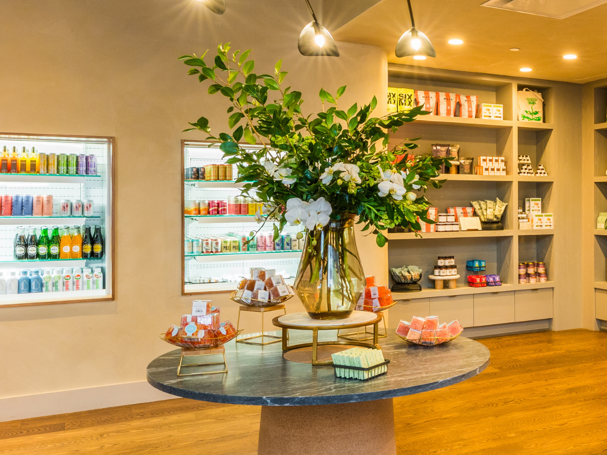Chic store interior with a round table, floral arrangement, and products on display; shelves stocked with beverages and goods.