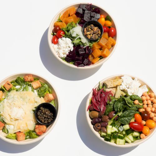 Three fresh salad bowls with mixed greens and veggies like beets, tomatoes, carrots, cucumbers, beans, and cheeses.