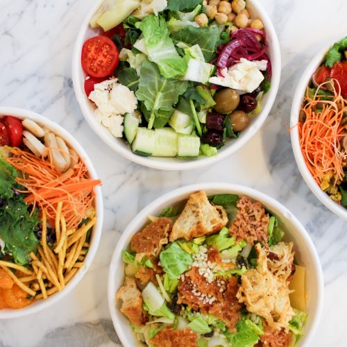 Four bowls of vibrant salads featuring fresh veggies, croutons, cheese, mixed greens, olives, and colorful garnishes on a marble surface.