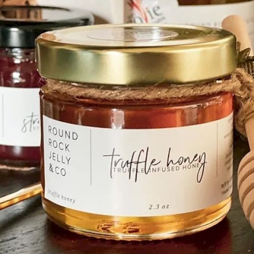 A jar of truffle-infused honey by Round Rock Jelly & Co with a gold lid, labeled as 2.3 oz, on a wooden surface.
