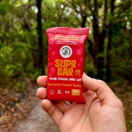 A hand is holding a Supr Bar energy snack labeled "Espresso Peanut Butter" in a red wrapper, with a forest path in the background.