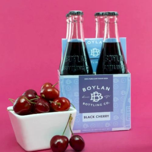 The image shows Boylan Bottling Co. Black Cherry soda in glass bottles, with a container of cherries beside it on a pink background.