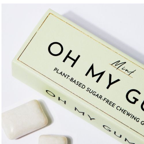 A box of mint-flavored, plant-based, sugar-free chewing gum labeled "OH MY GUM!" with a few pieces of gum beside it.