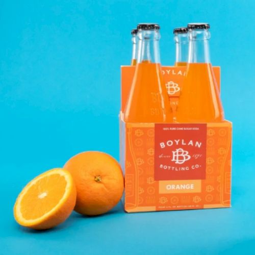 The image shows a pack of orange-flavored soda bottles labeled "Boylan Bottling Co." next to two whole oranges on a blue background.