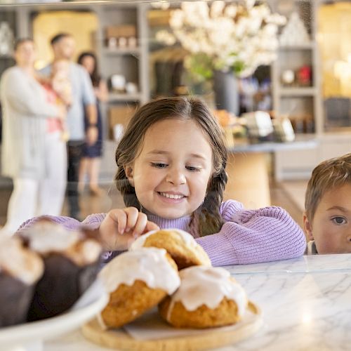 A girl and boy are looking at pastries inside a bakery, with more people in the background.