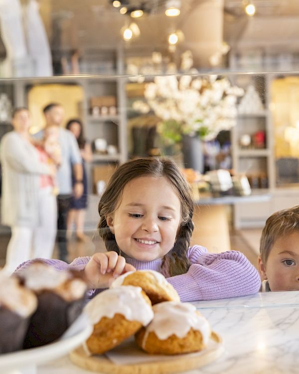 A girl and boy are looking at pastries inside a bakery, with more people in the background.