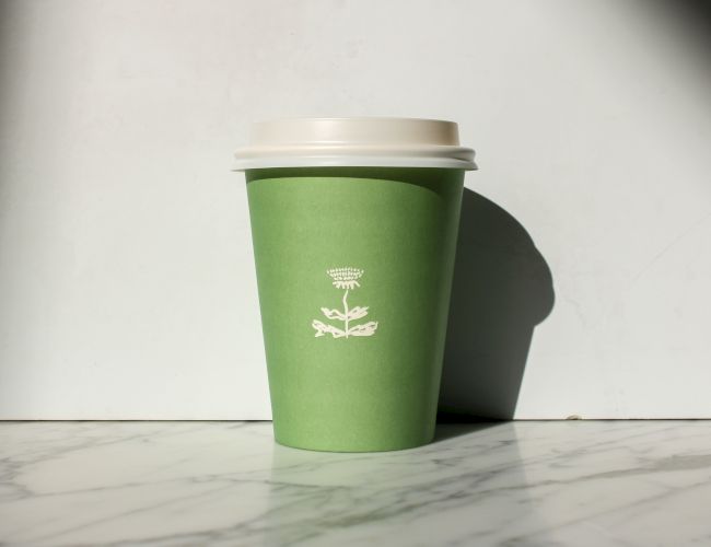 A green paper cup with a white lid sits on a marble surface, featuring a simple logo design in white.