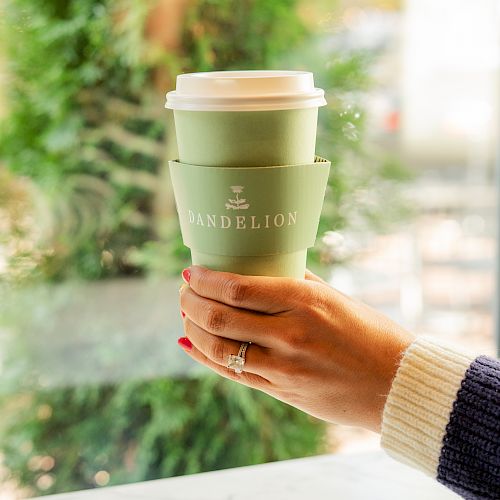 A hand holds a Dandelion branded coffee cup outdoors, with a green garden blur behind it, ready to sip.