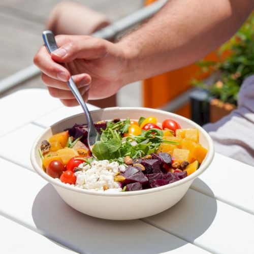 A person eats a colorful salad in the sun, mixing greens, tomatoes, beets, and cheese with a spoon.
