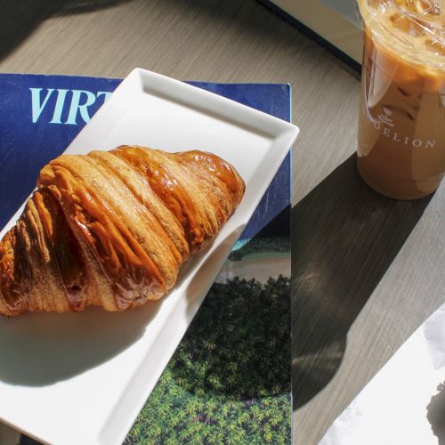 A toasted pastry on a white plate, an iced coffee drink, and an open booklet on a wooden table.