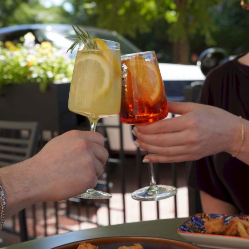 Two people clink tall cocktail glasses with lemon and rosemary on a sunny patio, sharing a toast outdoors.