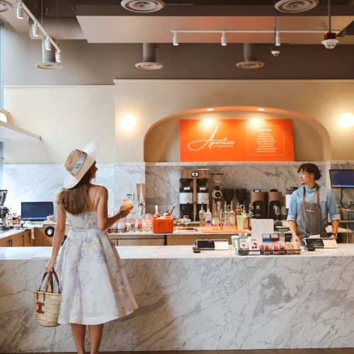 A woman in a sundress chats at a marble bar with drinks and a bartender in a modern cocktail lounge.