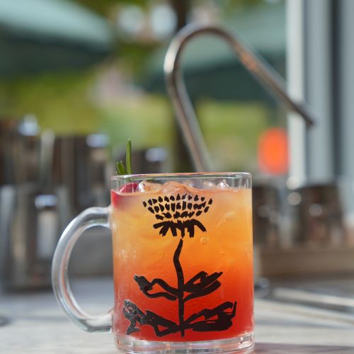 A glass mug of iced orange-red cocktail with a palm tree design, served outdoors under warm bokeh lights.