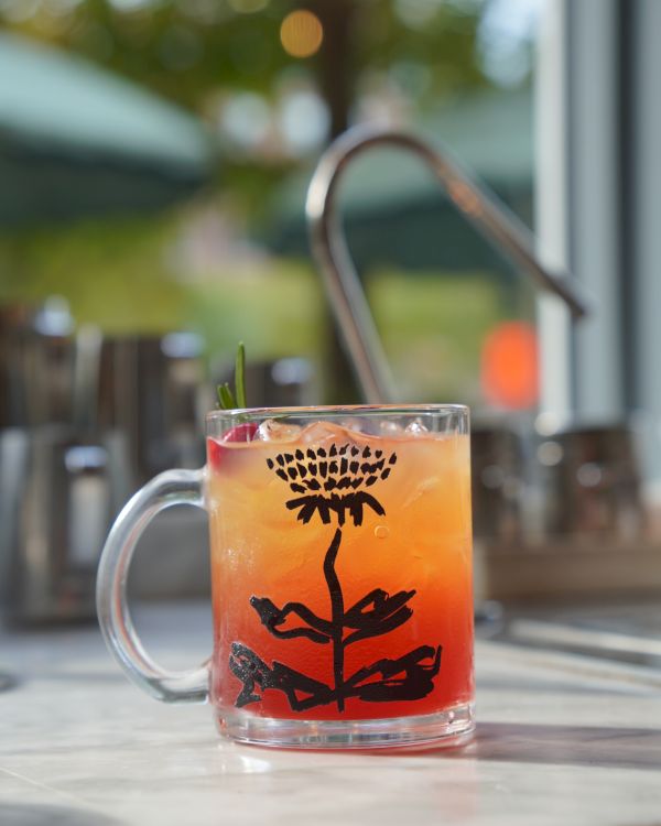 A glass mug of iced orange-red cocktail with a palm tree design, served outdoors under warm bokeh lights.