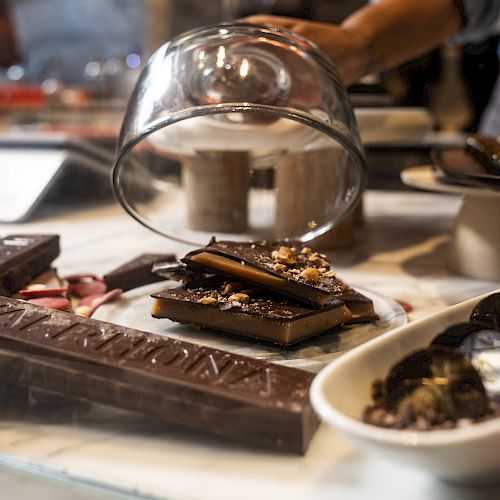 A pastry chef is sprinkling toppings over chopped chocolate on a tray while a bowl sits nearby, in a warmly lit kitchen.