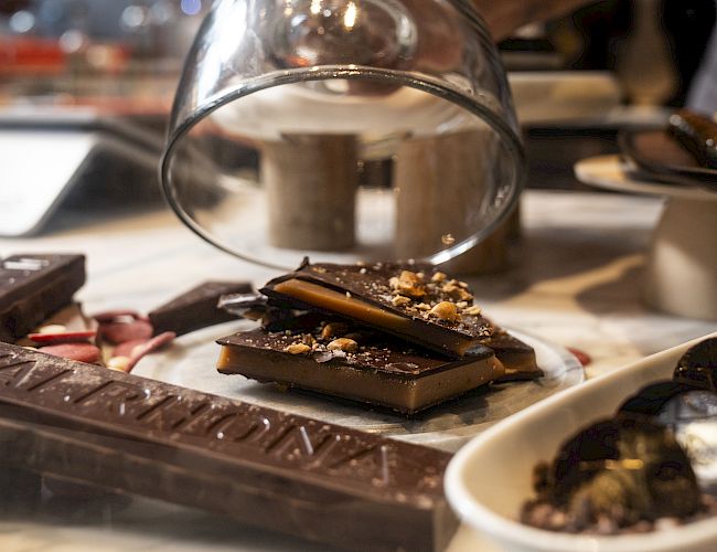 A pastry chef is sprinkling toppings over chopped chocolate on a tray while a bowl sits nearby, in a warmly lit kitchen.